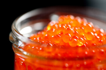 Red caviar in a glass jar, close up. Salmon caviar isolated on black background. Delicious gourmet...