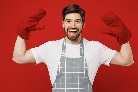 Young Cheerful Fun Happy Confident Caucasian Male Housewife Housekeeper Chef Cook Baker Man Wear Grey Apron Raise Up Hands In Oven Mittens Isolated On Plain Red Background Studio Cooking Food Concept