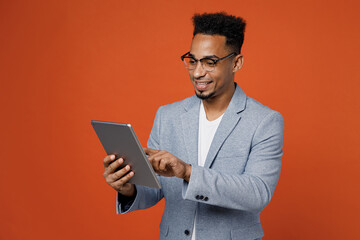 Young fun successful employee business man corporate lawyer wears classic formal grey suit shirt glasses work in office use digital tablet pc computer isolated on plain red orange background studio.