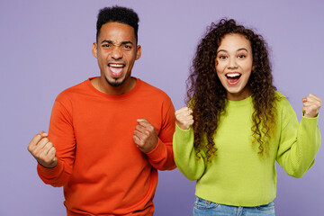 Young couple two friends family man woman of African American ethnicity wear casual clothes together do winner gesture celebrate clench fists say yes isolated on pastel plain light purple background.