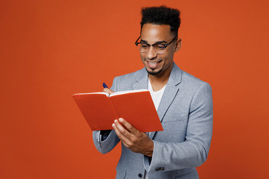 Young Smart Employee Business Man Corporate Lawyer Wear Classic Formal Grey Suit Shirt Glasses Work In Office Hold In Hands Diary Notebook Writing Notes Isolated On Plain Red Orange Background Studio.