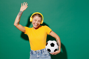 Young woman fan wear yellow t-shirt headphones cheer up support football sport team hold in hand soccer ball watch tv live stream listen music raise up hand isolated on dark green background studio. © ViDi Studio