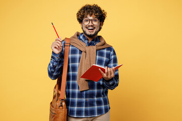 Young teen Indian boy IT student in casual clothes glasses bag write in notebook point finger pencil up with new idea isolated on plain yellow color background High school university college concept