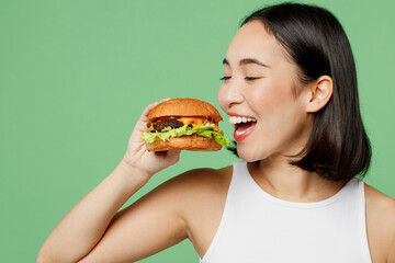 Close up young smiling happy fun woman wearing white clothes holding eating biting burger isolated on plain pastel light green background. Proper nutrition healthy fast food unhealthy choice concept.