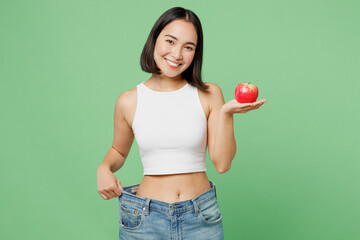 Young woman wears white clothes show loose pants on waist after weightloss hold red apple isolated on plain pastel light green background. Proper nutrition healthy fast food unhealthy choice concept.