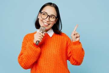 Young smiling woman of Asian ethnicity wear orange sweater glasses sing song in microphone point...