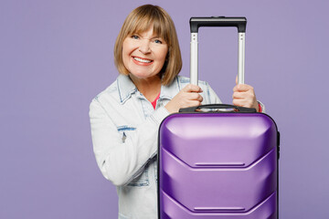 Traveler happy elderly woman 50s years old wear casual clothes hold suitcase isolated on plain pastel purple background. Tourist travel abroad in free spare time rest getaway. Air flight trip concept.