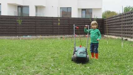 Funny little child with rubber boots work in garden with lawn mower, determined boy with big will, do not give up, young children give big lessons