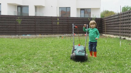 Funny little child with rubber boots work in garden with lawn mower, determined boy with big will,...
