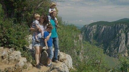 Naklejka na ściany i meble Parents holding children standing on top rock and look to river, mountain travel