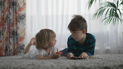 Portrait of little toddler and elder brother lying on carpet read a book, talk