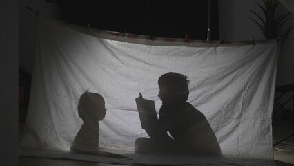 Big boy reading a tale to his smaller brother under a sheet tent with a lamp