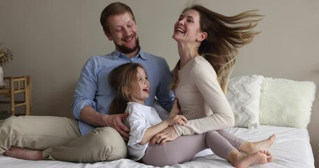 Young happy mother and father tickling cute little daughter sit on bed in bedroom, enjoy leisure at modern own house, spend carefree morning with beautiful child at home. Family ties, cherish, love