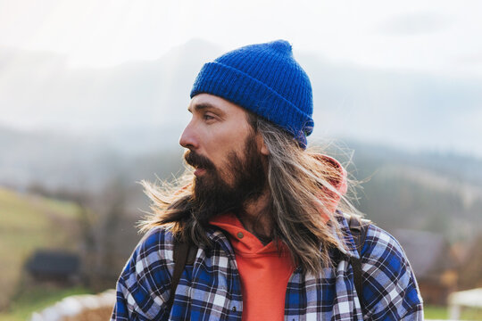 Portrait of hipster man with long hair wearing blue hat, sweatshirt and plaid jacket with mountain on the background while hiking.