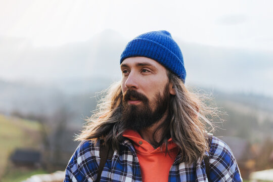 Man hiker staing with beautifule view on the mountains. Portrait of hipster man with long hair wearing blue hat and plaid jacket with mountain on the background.