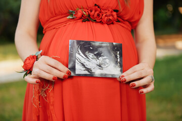 Pregnant woman in red dress holding ultrasound image above her belly.