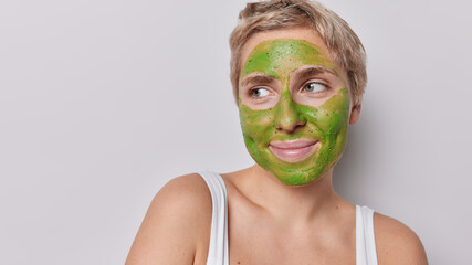 Close up shot of pleased short haired woman applies nourishing green mask for skin moisturising dressed in t shirt stands bare shoulders against grey studio background blank space for advertisement