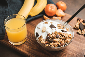 Granola cereal oatmeal with white yogurt, chocolate, banana fruit and nuts in a bowl on dark wooden board