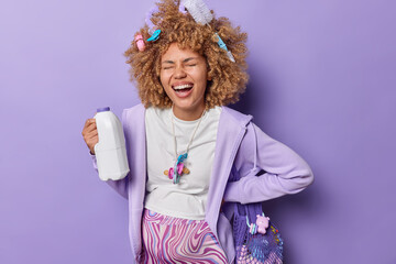 Horizontal shot of cheerful woman laughs positively going to give birth to baby carries nipples around neck holds bottle with milk exclaims loudly keeps eyes closed isolated over purple background.