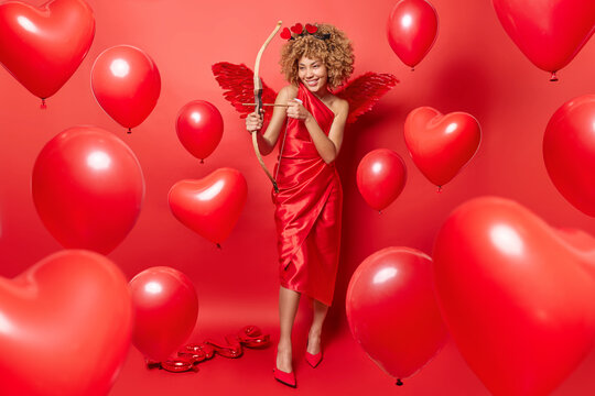 Positive woman with curly hair being in role of cupid shoots arrow wears long dress high heeled shoes and wings behind back isolated over red background heart shaped balloons around aims to find love