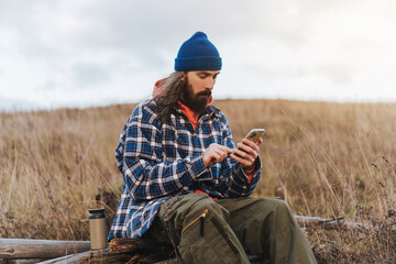 Young male travel photographer wearing blue beanie and jacket checking map on smartphone while a halt on the mountains on sunset.