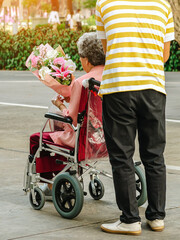 Back view of happy old senior woman sitting in a wheelchair and holding a bouquet in garden. Asian senior or elderly old lady woman holding beautiful flowers on wheelchair with caregiver son in park.