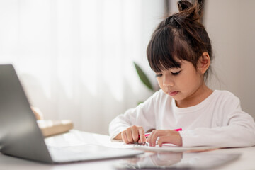 Fototapeta premium Asian schoolgirl doing her homework with laptop at home. Children use gadgets to study. Education and distance learning for kids. Homeschooling during quarantine. Stay at home