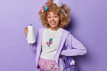 Horizontal shot of cheerful pregnant woman with big tummy holds plastic bottle with milk smiles toothily dressed in domestic clothes being at home at maternity leave isolated over purple background