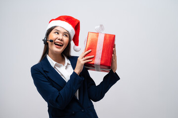 Beautiful Asian call center operator in Christmas hat talking with customer using headset and microphone holding gift box isolated on grey background.