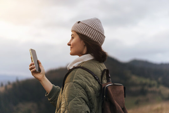 Pretty traveling woman standing on top of a mountain at sunset and using a mobile phone. Hiker female wearing beanie hat and taking pictures on the beautiful sunset in the mountains
