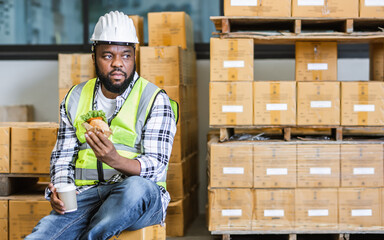African professional male worker, holding cup coffee, burger, having lunch, taking break, work in storage, warehouse or factory for delivery, wearing safety hat. Industry Business Concept.