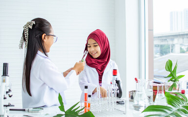 Two diverse multiethnic little girls wearing white ground uniform, hijab, headscarf, studying science in classroom at school, using stethoscope for research plants, happily smiling. Education Concept.