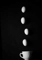 Black background. White cup with falling eggs in the middle of the cup.