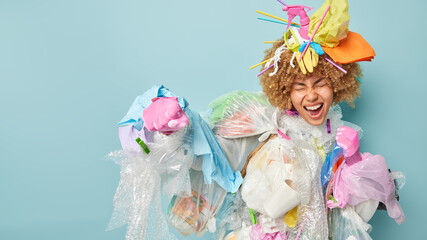 Positive curly haired female volunteer clenches fists pretends fighting against contamination asks not to pollute our planet makes costume from plastic garbage isolated on blue background blank space
