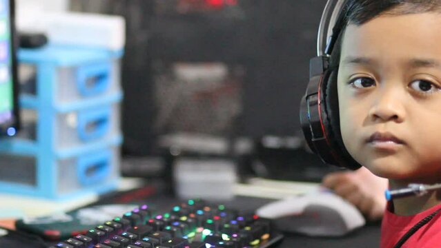 A little boy plays online games on a computer using a headset and a mechanical keyboard with colorful led lights.