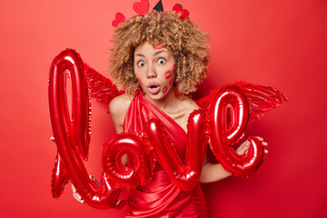 Surprised curly haired woman holds letter shaped balloons as symbol of love wears dress and wings...