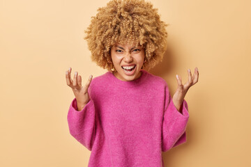 Angry curly haired woman shakes hands grimaces outraged expresses rage screams loudly dressed in pink jumper isolated over brown background stands full of hate being pressured and distressed