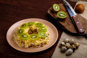 Layered salad with chicken and kiwi next to the board kiwi and quail eggs and a knife on a napkin.