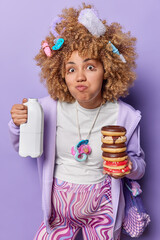 Funny curly haired young pregnant woman holds bottle of milk and pile of delicious sweet doughnuts dressed in casual clothes has addiction to sugar good appetite. Expectant mother with dessert