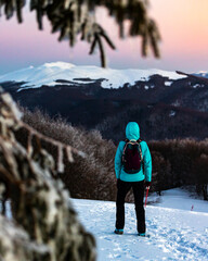 Backpacker girl admires colorful sunset while standing on top of a snowy mountain during cold winter weather