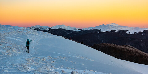 Backpacker girl admires colorful sunset while standing on top of a snowy mountain during cold winter weather