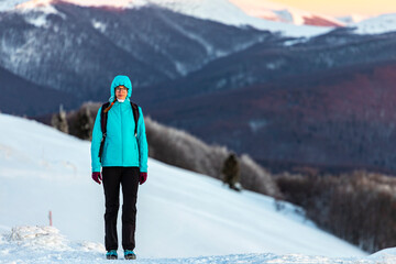 Backpacker girl walks across snowy ridge at sunset; hiking on snowy mountains during cold winter weather