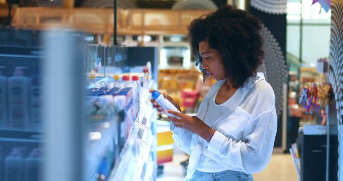 Young attractive dark-skinned woman with stylish afro hairstyle, standing in front of shelves with deodorant in the hygiene and personal care department of a supermarket, holding two and try to choose