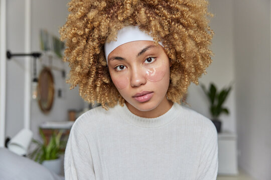 Indoor shot of serious curly haired woman wears headband and white jumper applies under eye hydrogel patches to remove dark circles and get rid of fine wrinkles poses against blurred home background
