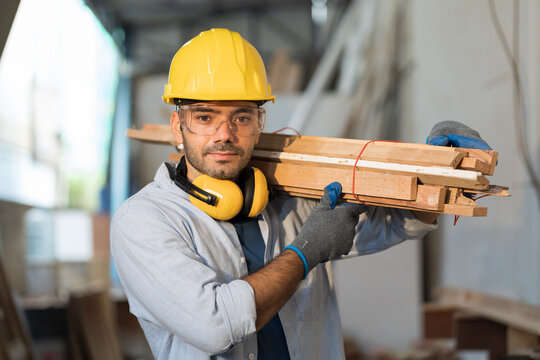 Male Carpenter Carrying Piece Of Wood On His Shoulder At Wood Processing Plants. Male Carpenter Working At Wood Workshop. Young Man Carpenter At Furniture Workshop