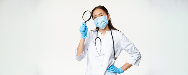 Image of asian female doctor, physician with magnifying glass, wearing medical mask and rubber...