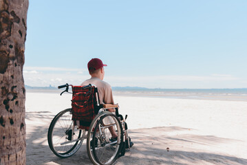 Fototapeta premium Back of young man with disability wearing a red cap on wheelchair, Activity outdoors on the beach background, Vacation on holiday and natural therapy and mental health concept.