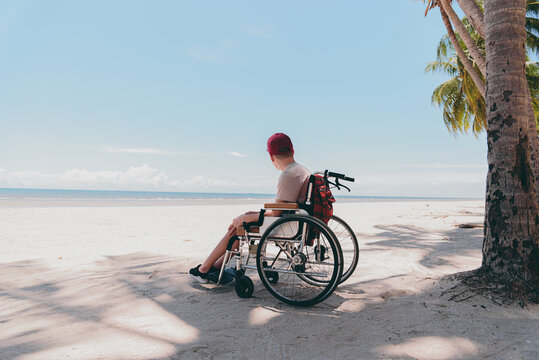 Back of young man with disability wearing a red cap  on wheelchair, Activity outdoors on the beach background, Vacation on holiday and natural therapy and mental health concept.