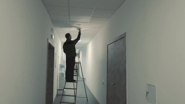 A master electrician is standing on a ladder and changing a light bulb in a dark corridor