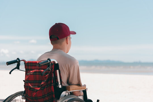 Back of young man with disability wearing a red cap  on wheelchair, Activity outdoors on the beach background, Vacation on holiday and natural therapy and mental health concept.